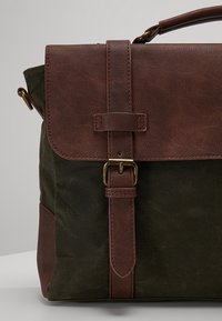 Rabatt en cuir marron avec une fermeture à boucle sur un corps en toile vert foncé ; comprend des accents en cuir marron et une couture robuste.
