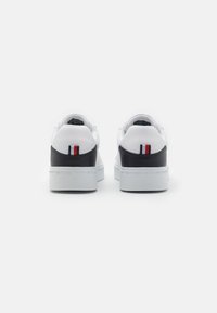 Zapatillas blancas con talón negro, con acentos texturizados y un pequeño detalle de bandera en rojo, blanco y azul. Suelas gruesas y blancas.