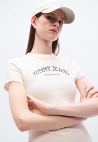 Ανοιχτό μπεζ εφαρμοστό t-shirt με κοντά μανίκια, με σκούρο μπλε κείμενο "TOMMY JEANS athletics". Φορεμένο με ανοιχτόχρωμο καπέλο.