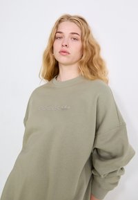 Olive krāsas lielizmēra sporta krekls, izgatavots no kokvilnas maisījuma, ar toņu izšūtu "adidas" logotipu un kritušiem plecu šuvēm.