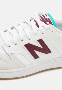 Weißer Sneaker mit maroonem "N"-Logo, Mesh- und Wildlederakzenten. Besitzt weiße Schnürsenkel, ein türkisfarbenes Zungendetail und eine strukturierte Gummisohle.