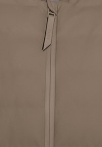 Hellbraune wasserdichte Jacke mit einem zentralen Reißverschluss und einem strukturierten Ziehband, das mit "RAINS" beschriftet ist. Durchgängige, glatte, matte Oberfläche.