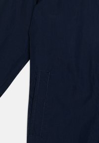 Navy blue jacket featuring a smooth texture, single side pocket with visible stitching, and an angled sleeve design. Lightweight material.