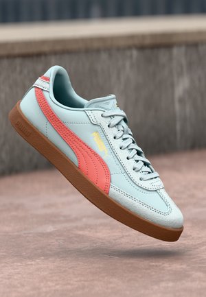 Jasnoniebieski sneaker Puma z koralowym paskiem i gumową podeszwą, pokazany w powietrzu na tle zewnętrznego betonu.