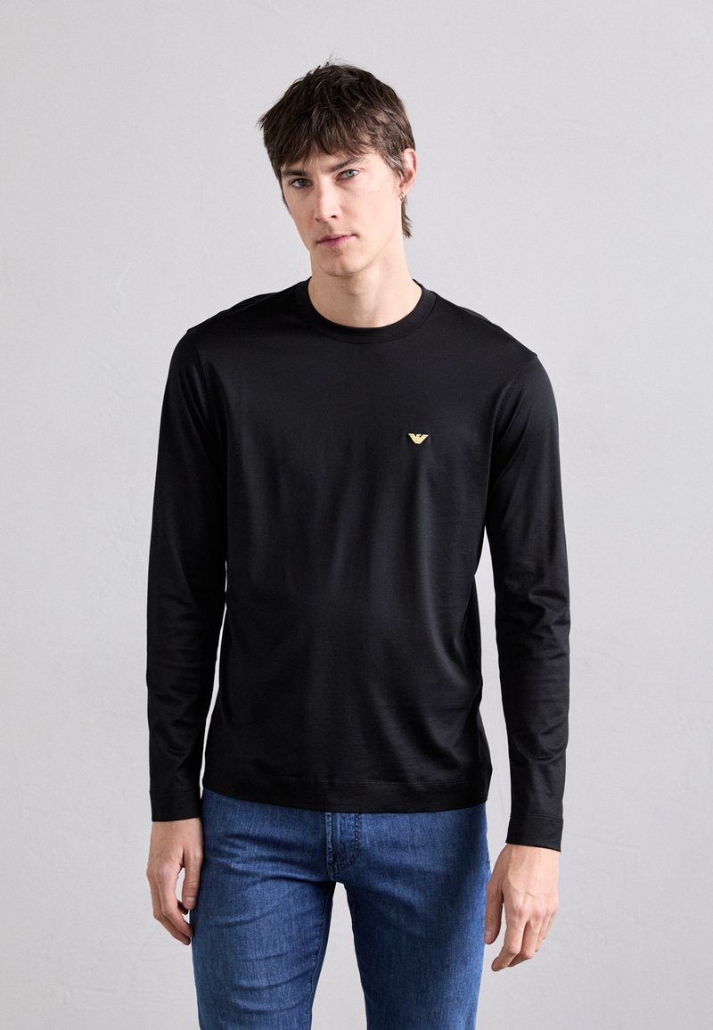 T-shirt noir à manches longues en tissu lisse, avec un petit logo doré sur la poitrine et un col rond. Porté avec un jean bleu.