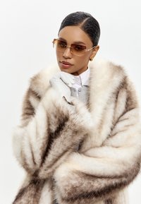 Cappotto in pelliccia sintetica nelle tonalità marrone e crema con un design voluminoso. Abbinato a guanti bianchi e occhiali da sole quadrati, creando un look stratificato.