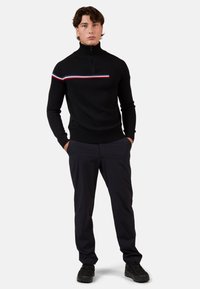 Schwarzer Strickpullover mit hohem Kragen und Reißverschluss, der über die Brust einen horizontalen Streifen in Rot, Weiß und Blau aufweist. Getragen mit dunklen Hosen.