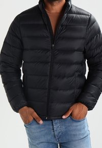 Giacca a vento nera con zip frontale, design trapuntato orizzontale e polsini elasticizzati, indossata sopra una camicia marrone e jeans blu.