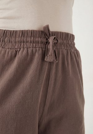 Zbliżenie osoby noszącej brązowe spodnie z elastycznym pasem i zawiązanym troczkiem oraz beżową bluzkę włożoną do środka.