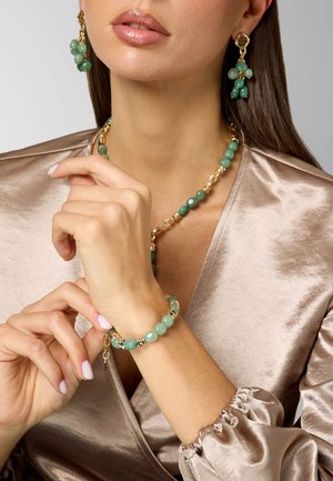 Donna che indossa un set di gioielli verdi di perline e oro, tra cui orecchini pendenti, collana e braccialetto, vestita con una maglia di raso beige lucido.