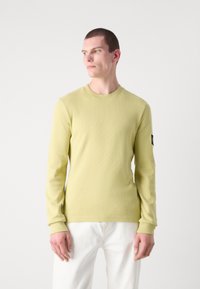 Langermet, tettsittende skjorte i lys grønn teksturert stoff med rund hals og ribbet mansjetter. Har et lite logo-merke på ermene.