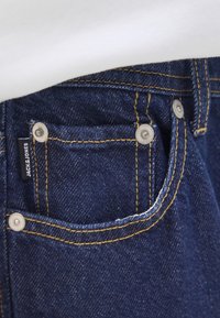 Gros plan sur une poche en denim bleu avec des détails en boutons argentés et une petite étiquette portant la mention « JACK & JONES ». L'arrière-plan est blanc.