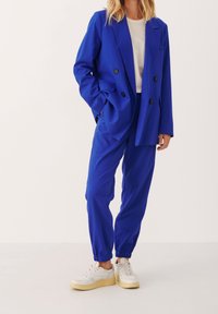 Kungligt blå dubbelknäppt blazer och matchande avsmalnande byxor i slät tyg, ihop med vita sneakers med tjock sula.