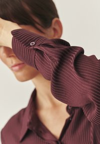 Burgundská prúžkovaná blúzka s manžetou na gombíky, jemnou textúrou a uvoľneným strihom. Tkanina sa elegantne splýva s jemným leskom.