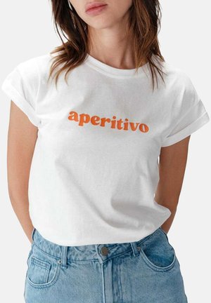 Femme portant un t-shirt blanc avec manches retroussées et texte orange "aperitivo" associé à un jean bleu clair taille haute.