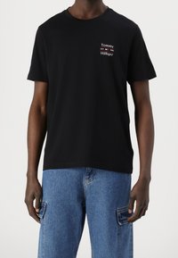 Mężczyzna w czarnym T-shircie Tommy Hilfiger z logo na lewej piersi oraz niebieskich bojówkach dżinsowych na jednolitym tle.