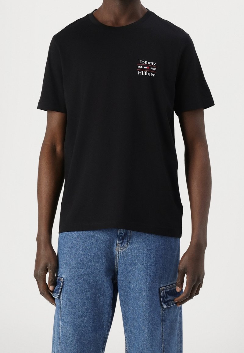 Mężczyzna w czarnym T-shircie Tommy Hilfiger z logo na lewej piersi oraz niebieskich bojówkach dżinsowych na jednolitym tle.