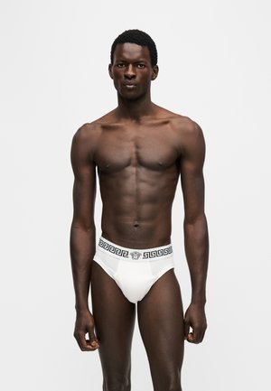Homme debout face à l’avant, portant un slip blanc avec une ceinture noire à motifs géométriques, sur un fond uni clair.
