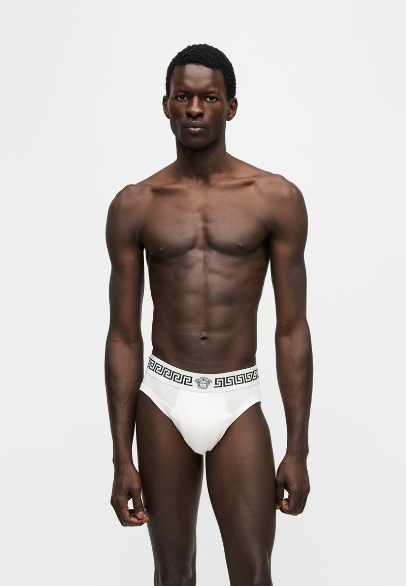 Homme debout face à l’avant, portant un slip blanc avec une ceinture noire à motifs géométriques, sur un fond uni clair.