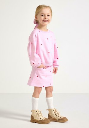 Smilende ung pige iført en lyserød sweatshirt og nederdel med røde hjerter, hvide knæstrømper, tanfarvede snørebåndsstøvler og et lyserødt hårelastik.