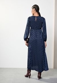 Marineblaues, langärmeliges Kleid mit gepunktetem Muster, geraffter Taille und einem Schlüsselloch-Detail am Rücken. Kombiniert mit braunen Stiefeletten und einer Clutch.