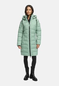 Kvinna stående i en ljusgrön lång vadderad jacka med huva, svarta byxor och svarta ankelstövlar på en vit bakgrund.