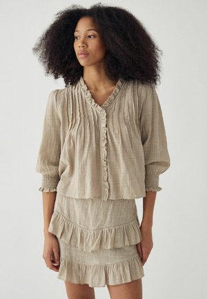 Nő dús göndör hajjal, bézs, texturált blúzt visel mélyített varrásokkal és hozzá illő fodros szoknyával, egy egyszerű háttér előtt áll.