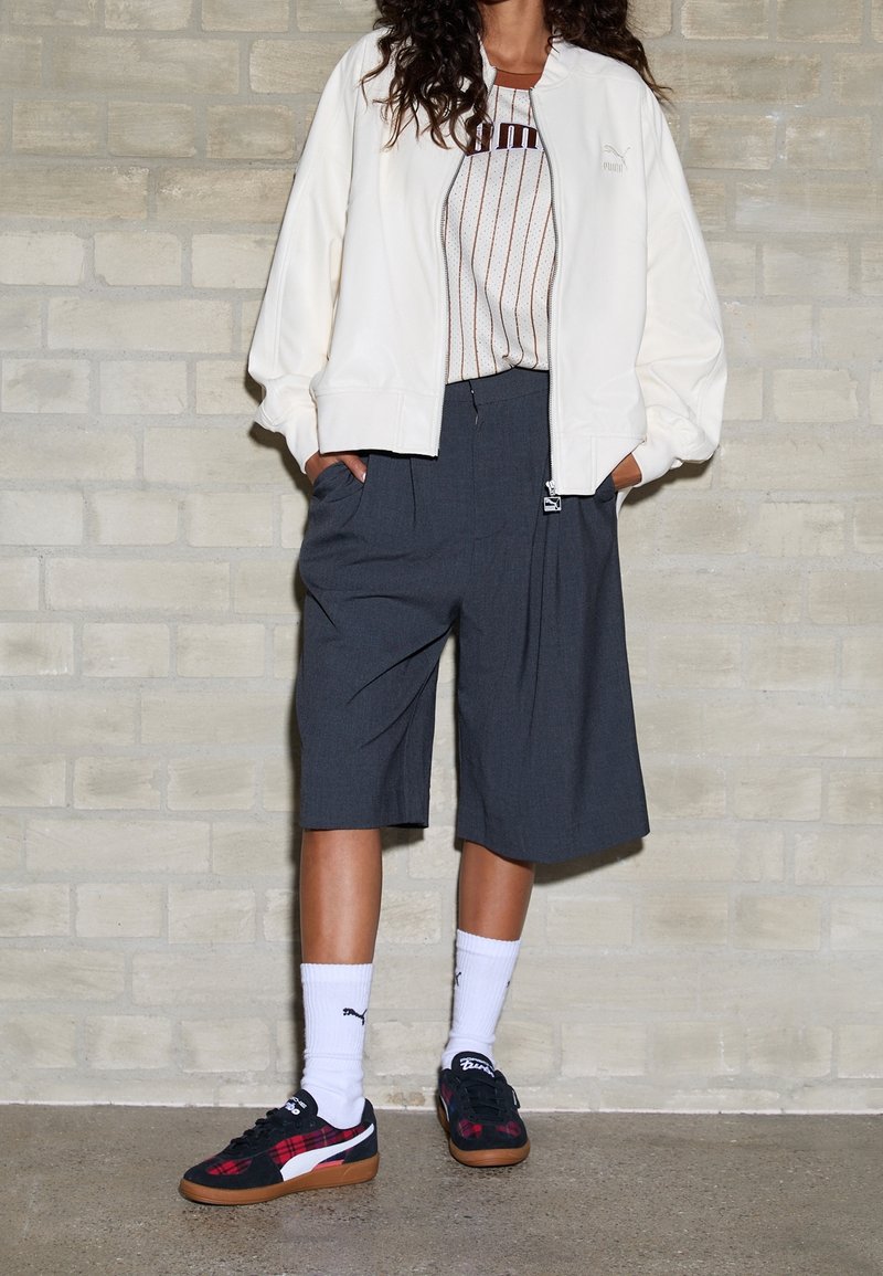 Giacca bomber bianca, top a maniche corte a righe, pantaloni corti grigi sartoriali e sneakers nere a scacchi con suola color gomma, abbinate a calze bianche.