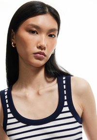 Gestreepte tanktop in marineblauw en wit, met geribbelde textuur, brede halslijn en dikke bandjes. Model draagt creolen.