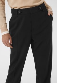 Pantalons noirs ajustés à taille haute, comportant deux poches avant, une texture lisse et des boutons hexagonaux distinctifs à la taille.