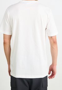 Osoba oblékající jednoduché bílé tričko s krátkým rukávem a tmavé cargo šortky, stojící zády k fotoaparátu na bílém pozadí.