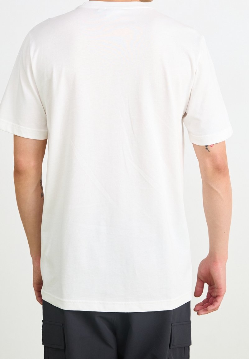 Osoba oblékající jednoduché bílé tričko s krátkým rukávem a tmavé cargo šortky, stojící zády k fotoaparátu na bílém pozadí.