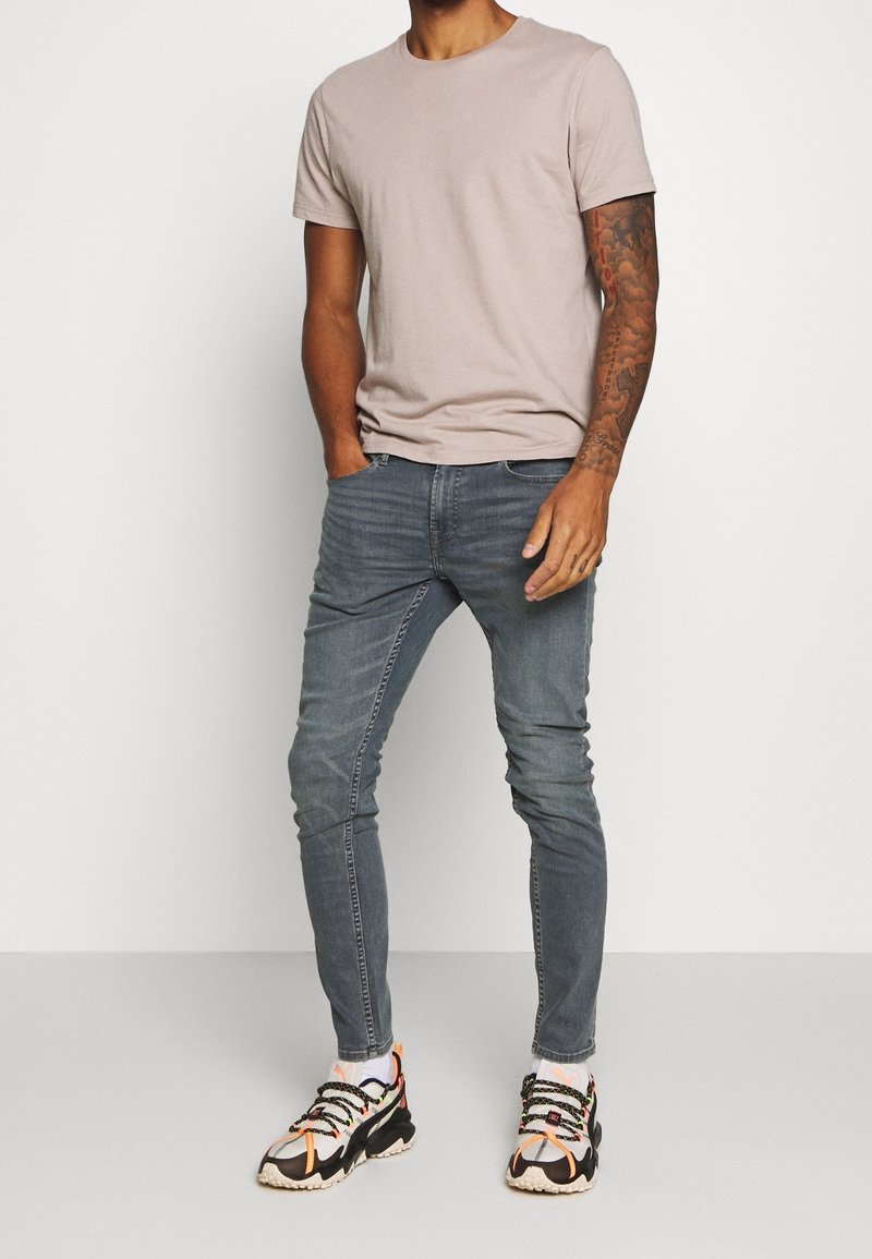 Homme portant un t-shirt beige clair à manches courtes, un jean gris slim, des chaussettes blanches et des chaussures de sport noires et orange, debout sur fond blanc.