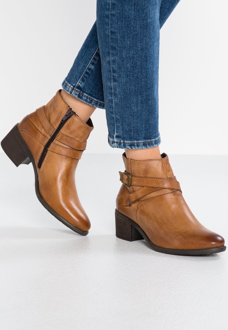 Bottes basses en cuir camel avec un petit talon, des sangles décoratives et une fermeture éclair latérale, présentées avec un jean bleu délavé sur fond blanc.