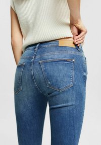 Mörkblå denimjeans med en figursydd design, utrustad med två bakfickor och en ljusbrun läderetikett vid midjan. Tyget har en lätt textur.