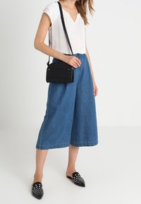 Kvinna i vit kortärmad blus, vidblanka blå denim culotter, svarta spikade slip-on skor och svart axelremsväska.