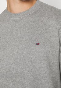 Szara bawełniana bluza z okrągłym dekoltem, o subtelnej fakturze, z małym haftowanym logo na piersi, zawierającym czerwone, białe i niebieskie akcenty.