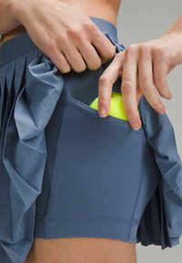 Fållad blå atletisk kjol med ficka, där en hand lägger en gul tennisboll i fickan, vilket framhäver den mjuka tygstrukturen.