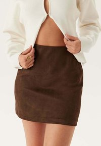 Jupe mini en suède marron avec une texture lisse, une forme droite et un ourlet simple. Associée à un haut couleur crème avec un décolleté en trou de serrure.
