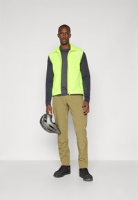 Bright green sleeveless jacket over a dark gray long-sleeve shirt, paired with light brown pants and black shoes; holding a gray helmet.