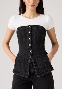 Črn denim korzet z navpičnim šivom, belimi gumbi in stranskimi žepi, v kombinaciji s prilegajočim belim rebrastim majico.
