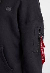 Manche de sweat-shirt noire avec une poche zippée et un porte-clé en tissu rouge portant l'inscription "REMOVE BEFORE FLIGHT."