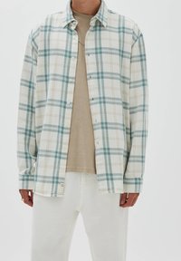 Chemise en flanelle à carreaux crème, bleu sarcelle et beige ; dotée de boutons à l'avant, d'un col et de manches longues. Portée sur un t-shirt beige et un pantalon blanc.