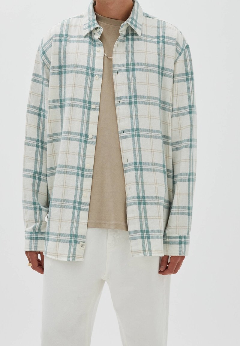 Chemise en flanelle à carreaux crème, bleu sarcelle et beige ; dotée de boutons à l'avant, d'un col et de manches longues. Portée sur un t-shirt beige et un pantalon blanc.