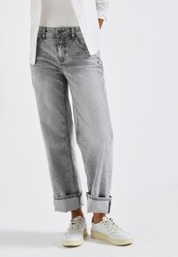 Persona che indossa jeans grigi chiari con risvolti, sneakers bianche, un top bianco e una camicia bianca abbottonata, con le mani nelle tasche su uno sfondo bianco.