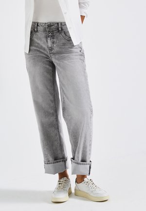 Person in hellgrauen, hochgekrempelten Jeans, weißen Sneakers, weißem Oberteil und weißem, knöpfbarem Hemd mit Händen in den Taschen vor weißem Hintergrund.