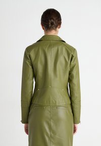 Femme avec les cheveux en chignon portant une veste en cuir ajustée olive et une jupe assortie, vue de dos sur fond blanc.