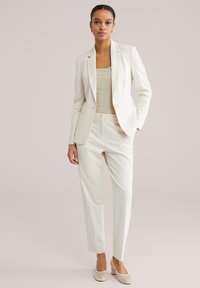 Moteris dėvi kreminės spalvos pritaikytą švarką, atitinkančias kelnes, smėlio spalvos ribuotos faktūros viršų ir kreminės spalvos batelius su blokiniu kulnu neutraliame fone.