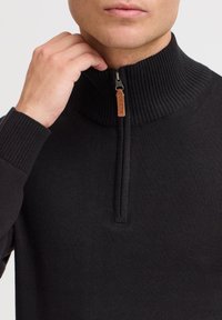 Pull en maille noire avec un col montant et une fermeture éclair mi-hauteur, doté de bords-côtes au niveau des poignets et du col, ainsi qu'un détail de tirette en cuir.