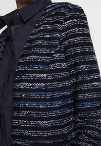 Gros plan sur une veste texturée bleu marine avec des rayures horizontales argentées et bleues sur une chemise col bleu foncé.